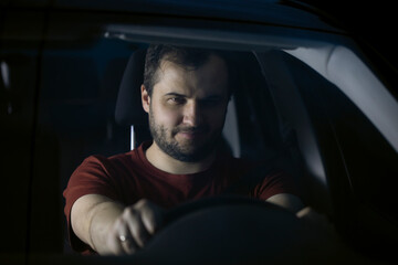 Unshaven confident serious young smiling millenial driver sits at steering wheel of car at night thoughtfully looking ahead. safe driving concept. Loneliness Concept.