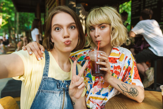 Image of women taking selfie and gesturing peace sign while drinking soda
