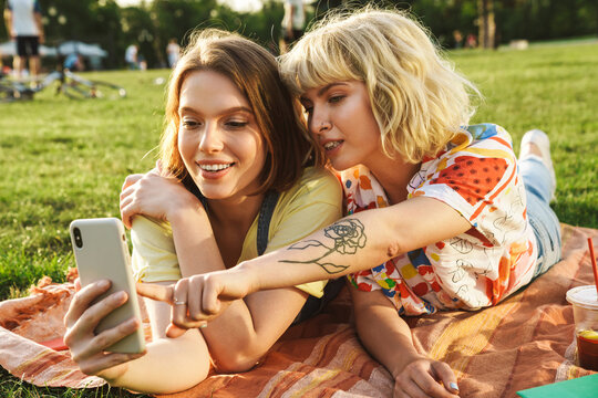 Image of pleased nice two women using cellphone while have picnic