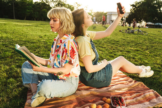 Image of women using cellphone and reading book while resting on grass