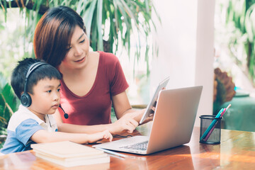 Young asian mother and son using laptop computer and tablet for study and learn together at home, boy wearing headphone for e-learning with distancing, teacher or mom support child, education concept.