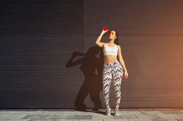 Young fit woman with beautiful figure refreshing with energy drink against black wall in the city,female runner taking break after workout standing against background with her amazing feminine shadow