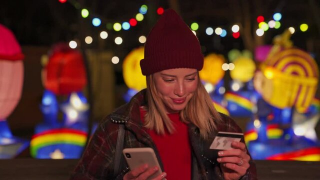 Smiling young blonde girl wearing autumn clothes paying the bills with smartphone and credit card over neon lighting background