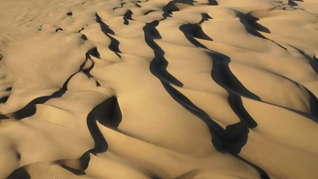 4K bird-eye view on the amazing wavy shape and pattern of sand hills. Aerial view of a drone flying over the scenic landscape of massive golden dunes at sunset. Trip to the wild desert nature, USA