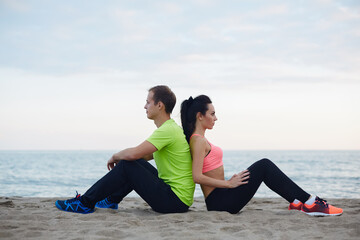 Fototapeta na wymiar Male and female runner sitting on the sand enjoying beautiful evening, young fit couple having break after workout outdoors, beautiful couple of athletes resting after run sitting on seashore