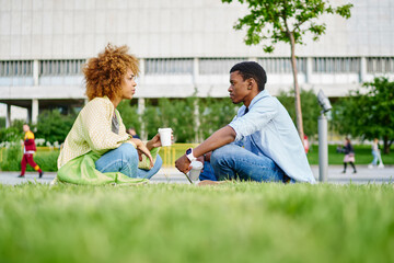 African american hipster girl sitting against boyfriend and communicating about relationship.Two friends talking with each other and drinking tasty coffee sitting on green grass in park