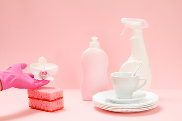Woman's hand in nitrile glove with an orchid flower, bottle of dishwashing liquid and utensil on a pink background.