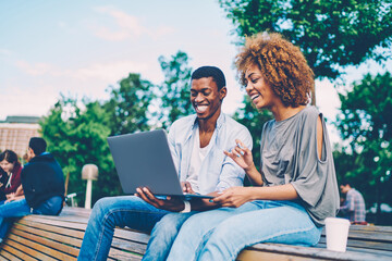 Cheerful afro american friends having fun watching movie together in city park satisfied with good internet connection, happy couple in love enjoying date on spring sunny day blogging in networks