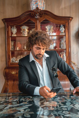 Street style an elegant black suit, white shirt and sunglasses. With a handsome young caucasian man with long hair in the city. Fashion editorial. In a hotel sitting sending a message on the phone