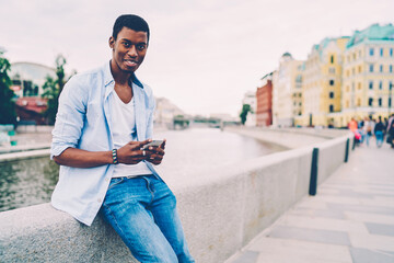 Half length portrait of smiling afro american hipster guy sending message via email on smartphone during strolling outdoors.Young positive tourist looking at camera while spending leisure time