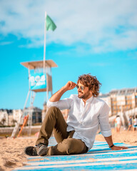 Summer lifestyle with a young dark-haired Caucasian man with long beard, sunglasses and a white shirt on a beach. San Sebastian, Basque Country. Sitting looking out to sea next to a beach watcher post