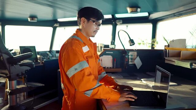Filipino deck Officer on bridge of vessel or ship wearing coverall during navigaton watch at sea . He is using laptop, electronic paperwork at sea, concept of reporting