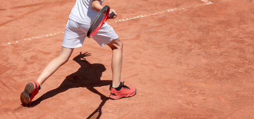 Boy plays tennis on red clay court. Child on tennis court hits forehand. Young tennis player with racket in action. Kids sports background with shadow. Banner size. Copy space
