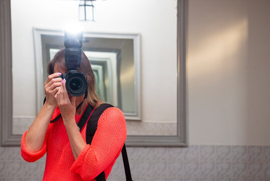 Woman with a camera in a bright pink sweater taking photos of her reflection in a mirror corridor