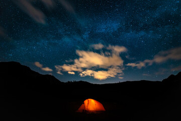 Orange tent with a burning light inside stands in a clearing under the starry sky. On the horizon mountains and forest. Concept of a beautiful starry night sky and the Milky Way. Night in the mountain