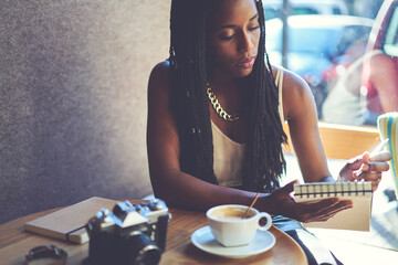 Pensive creative writer enjoying working process in coffee shop making researchers for startup project, talented afro american female author creating article in notepad analyzing information