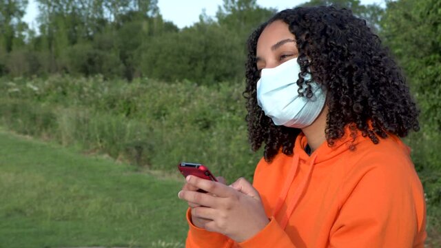 Beautiful mixed race African American girl teenager young woman wearing a face mask during COVID-19 Coronavirus pandemic using her smartphone or cell phone outdoors for social media 