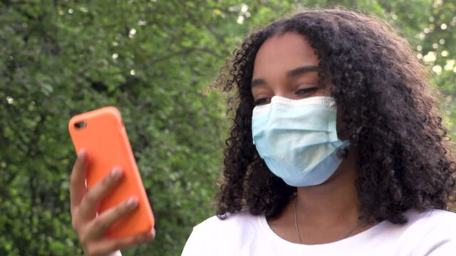 Beautiful mixed race African American girl teenager young woman wearing a face mask during COVID-19 Coronavirus pandemic using her smartphone or cell phone outdoors for social media 