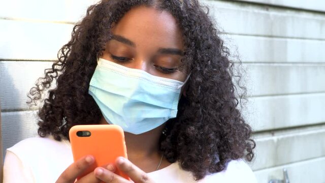 Beautiful mixed race African American girl teenager young woman wearing a face mask during COVID-19 Coronavirus pandemic using her smartphone or cell phone for social media 