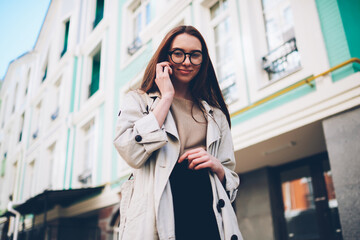 Half length portrait of positive gorgeous female in stylish spectacles talking on phone using mobile application during walking at street.Young hipster girl looking at camera while standing outdoors