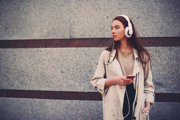 Charming pensive female music lover dressed in trendy casual outfit enjoying favorite songs waiting for meeting outdoors, teenage girl listening composition from playlist standing on advertising area