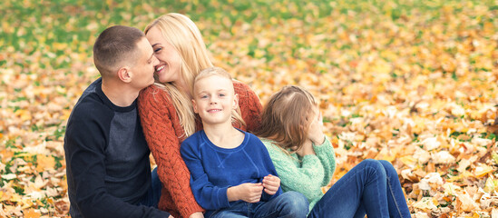 Fototapeta premium Portrait of young family sitting in autumn leaves. Parents kissing and sitting with children in the autumn park