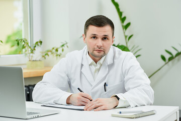 Fototapeta premium A caucasian doctor in a white lab coat is filling out a patient card with a diagnosis and listening in a hospital. A therapist near a laptop is waiting for a patient in a doctor's office.