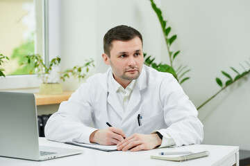 Fototapeta premium A caucasian doctor in a white lab coat is filling out a patient card with a diagnosis and thinking in a hospital. A therapist near a laptop is waiting for a patient in a doctor's office.