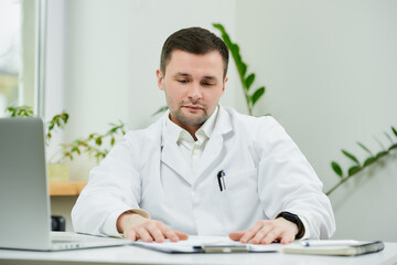A serious caucasian doctor in a white lab coat is sitting relaxed at his desk and looking at...