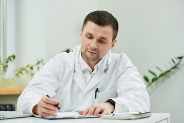 A serious caucasian doctor in a white lab coat is sitting at his desk and carefully examining the patient’s medical history in a hospital. A therapist near a laptop in a doctor's office.