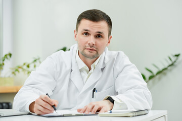 A serious caucasian doctor in a white lab coat is sitting at his desk and distracted from examining...