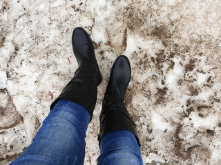 Women's feet in black boots on the dirty snow.