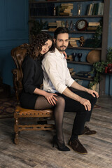 Obraz premium Vertical portrait of young couple sits in an unusual pose on chair and hugs tenderly. Brunette husband and wife in the old antique appartment. Mr and mrs in formal suits are posing. Vogue and luxury.