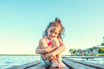 Fototapeta na wymiar little happy girl with curly hair sits on a wooden pier in the summer on the lake on a background of blue water. Looks into the camera lens