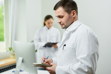 A caucasian doctor with a notebook and a pen records the patient’s medical history in a hospital. A female therapist is doing notes in a clipboard near an all in one computer in a clinic.