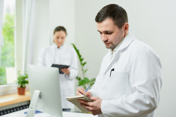 A caucasian doctor with a notebook and a pen is thinking about the patient’s medical history in a hospital. A female therapist is doing notes in a clipboard near an all in one computer in a clinic.