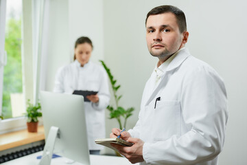 A serious doctor with a notebook and a pen is thinking about the patient’s medical history in a hospital. A female therapist is doing notes in a clipboard near an all in one computer in a clinic.