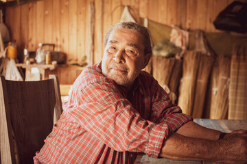 Old artisan in a local community of Garopaba, Brazil. Craftsman resting at his wooden workshop. Elder wearing red plaid shirt