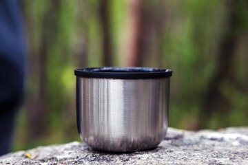 A mug from a thermos is standing in the woods on a stone.