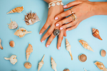 beautiful woman hands with pink manicure holding sea shells, lot of rings on fingers on blue background, luxury jewelry concept
