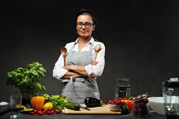 Female chef posing next to a table in kitchen. Studio photo on a dark background