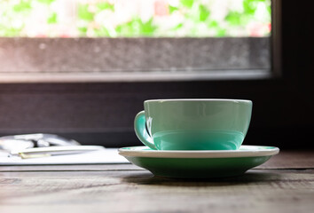 Close up white coffee cup with sunglasses and a pen on black wood table near window with light shade on tabletop at cafe.