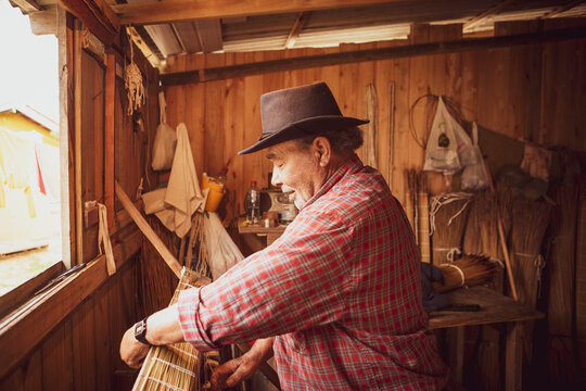 Traditional handicraft mat made of dry reed in a local community of Garopaba, Brazil. Artisan working at his wooden workshop. Craftsman wearing red plaid shirt and brown hat