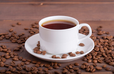 Cup of tasty black boiled coffee on a wooden background. A cup stands on a plate, roasted coffee beans are scattered around.