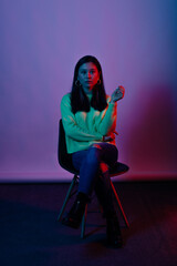 Vertical portrait of a young woman sitting on c chair in studio with red and blue light over white background.