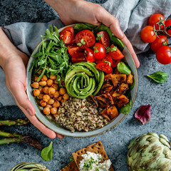 Hands holding healthy superbowl or Buddha bowl with salad, sweet potatoes, chickpeas, quinoa, tomatoes, arugula, avocado on light blue background. Healthy vegan food, clean eating, top view