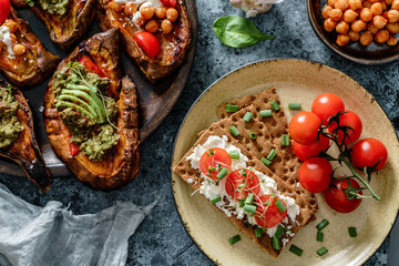 Baked sweet potato toast with roasted chickpeas, tomatoes, goat cheese, sauce guacamole, avocado, seedlings on wooden board over blue background. Healthy vegan food, clean eating, dieting, top view