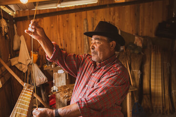 Traditional handicraft mat made of dry reed in a local community of Garopaba, Brazil. Artisan working at his wooden workshop. Craftsman wearing red plaid shirt and brown hat