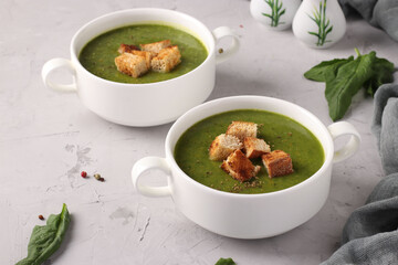 Green spinach puree soup served with croutons in two white bowls on a gray concrete background