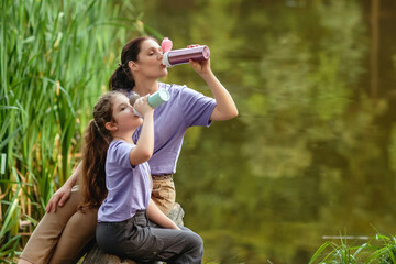 Mother and daughter on a walk in the park drinking from vacuum flask. Concept of outdoor recreation using reusable dishes. A bottle for coffee or tea to go.